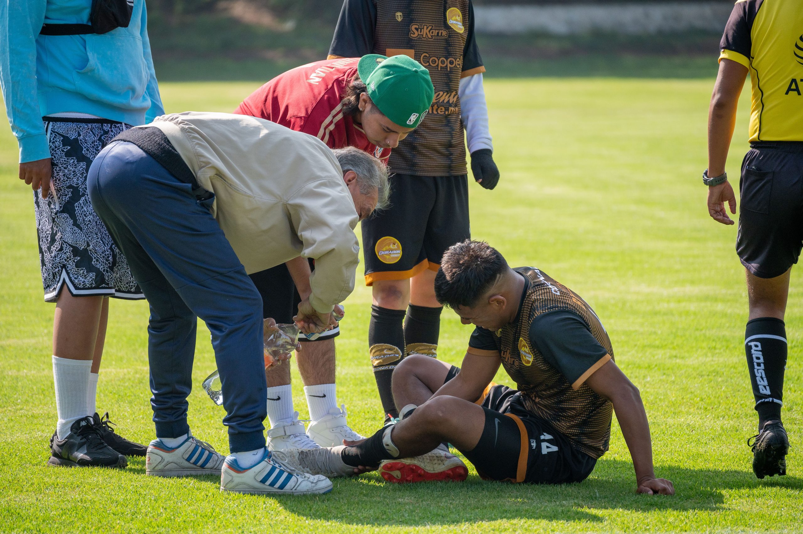Footballeur assis sur la pelouse soigné à la cheville par un staff penché, coéquipiers et arbitre autour.