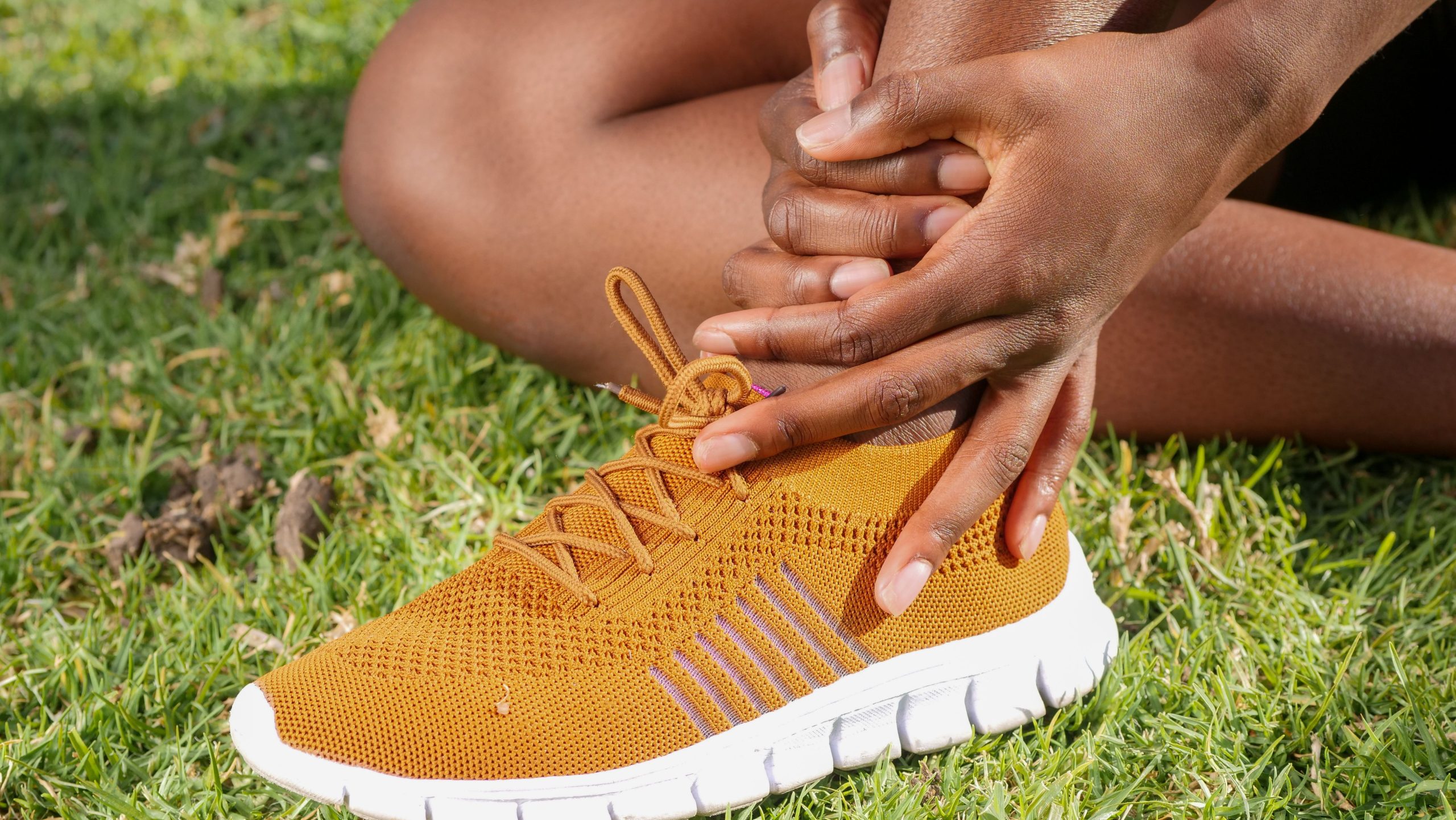 Coureuse assise dans l'herbe tenant son talon douloureux au-dessus d'une basket orange, tendon d'Achille sensible.