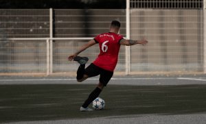 Footballeur en maillot rouge numero 6 s'elancant pour frapper un ballon sur un terrain synthetique.