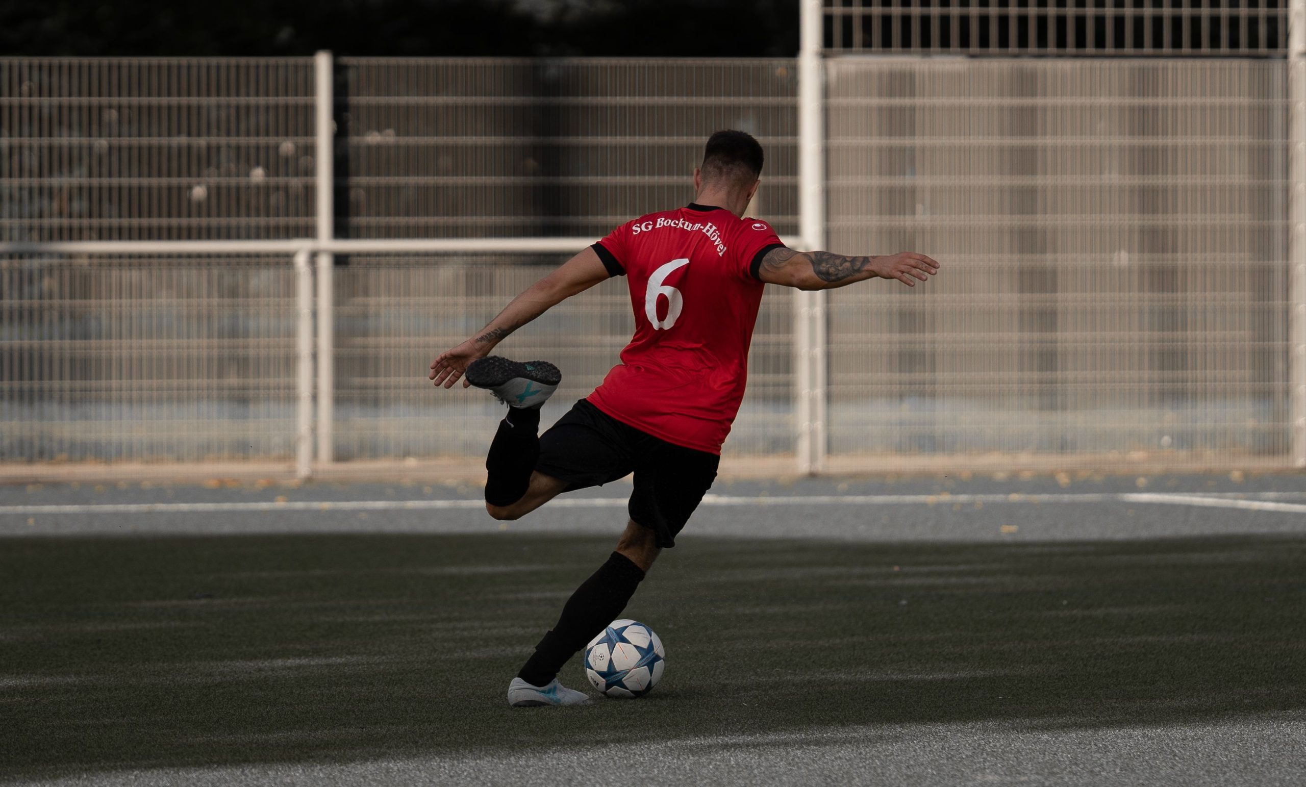 Footballeur en maillot rouge numero 6 s'elancant pour frapper un ballon sur un terrain synthetique.