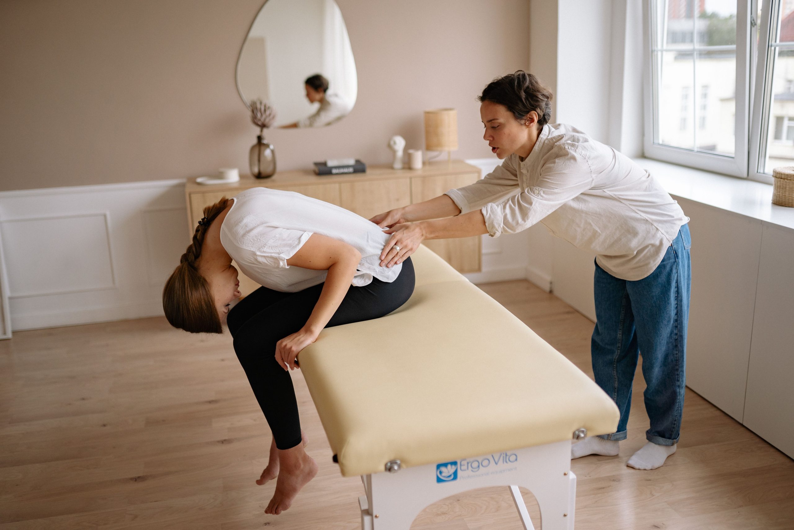 Kinesitherapeute palpant le bas du dos d'une patiente penchee en avant sur une table de soins.