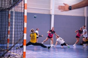 Gardienne de handball en plongeon devant son but, ballon en vol et joueuses en duel sur parquet de gymnase.