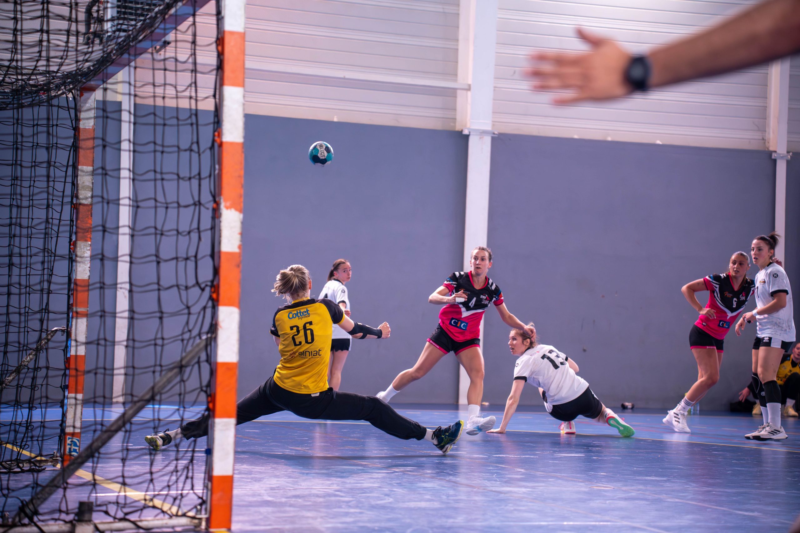 Gardienne de handball en plongeon devant son but, ballon en vol et joueuses en duel sur parquet de gymnase.
