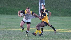 Joueur de rugby ballon sous le bras esquivant un plaquage adverse lors d'un match sur pelouse.