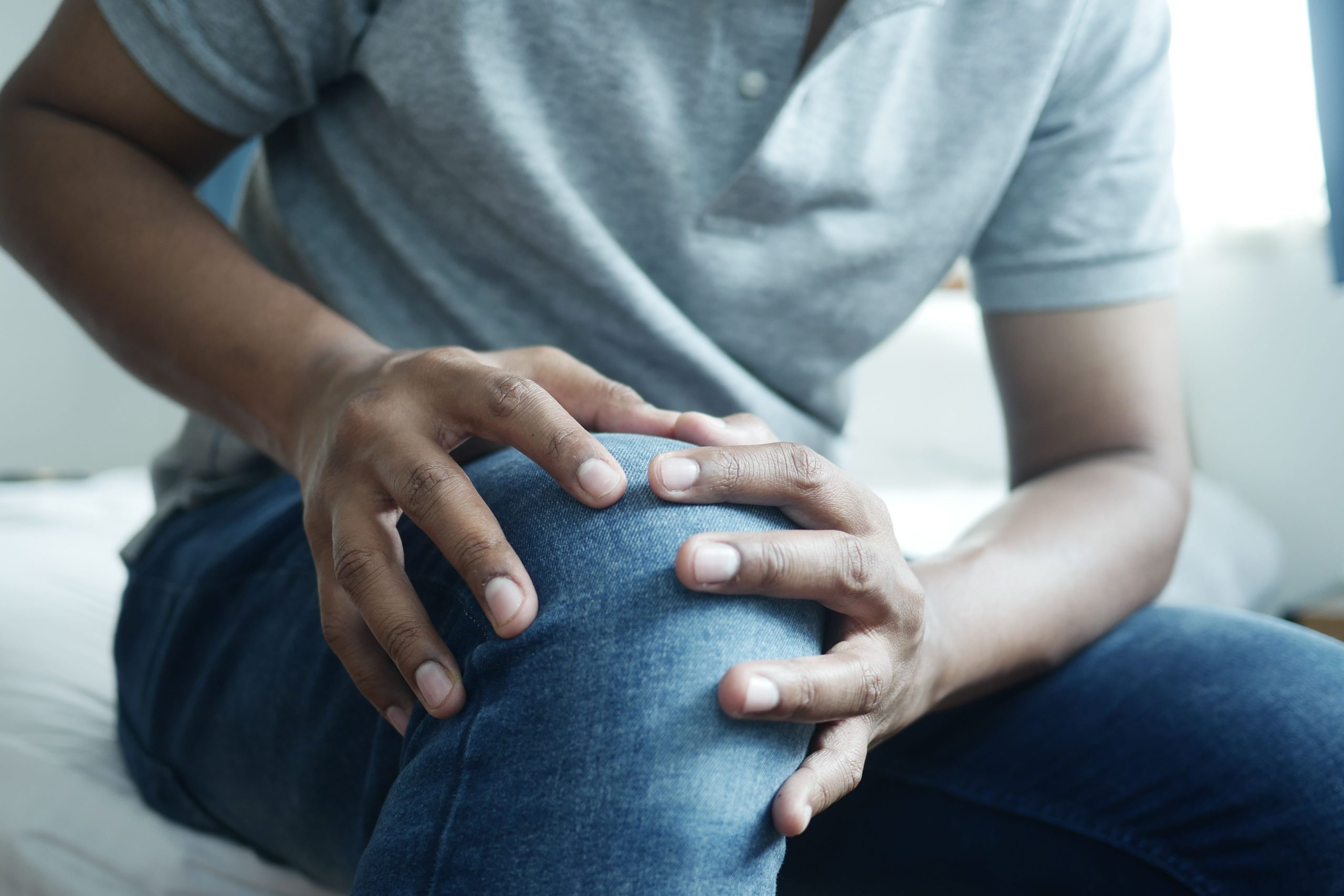 Homme assis tenant son genou douloureux a deux mains a travers son jean, evoquant une gene articulaire post-traumatique.