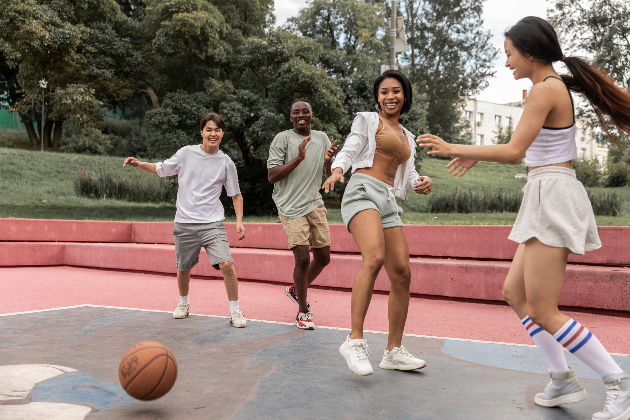 Quatre jeunes adultes souriants courent vers un ballon de basket sur un playground extérieur.