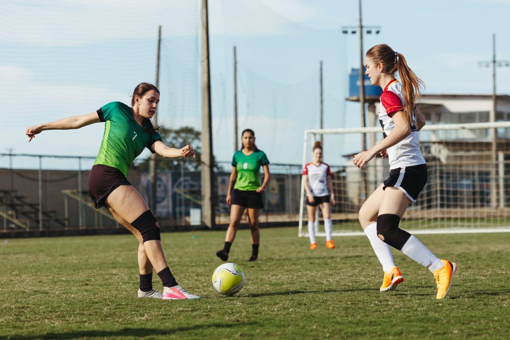 Footballeuses en duel pour le ballon sur un terrain, l'une portant une genouillere de soutien apres contracture.